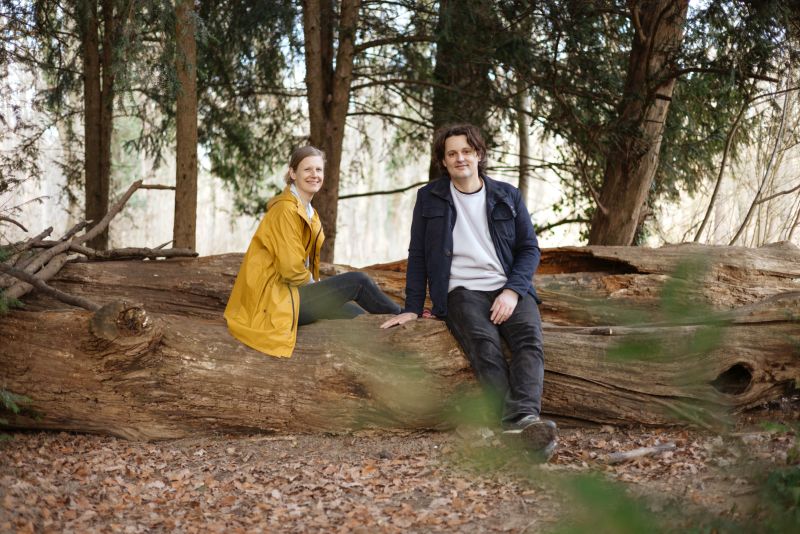 Ganzfigur: Carolin Lange und Dico Kruijsse sitzen auf einem Baumstamm im Wald und  schauen in die Kamera. 