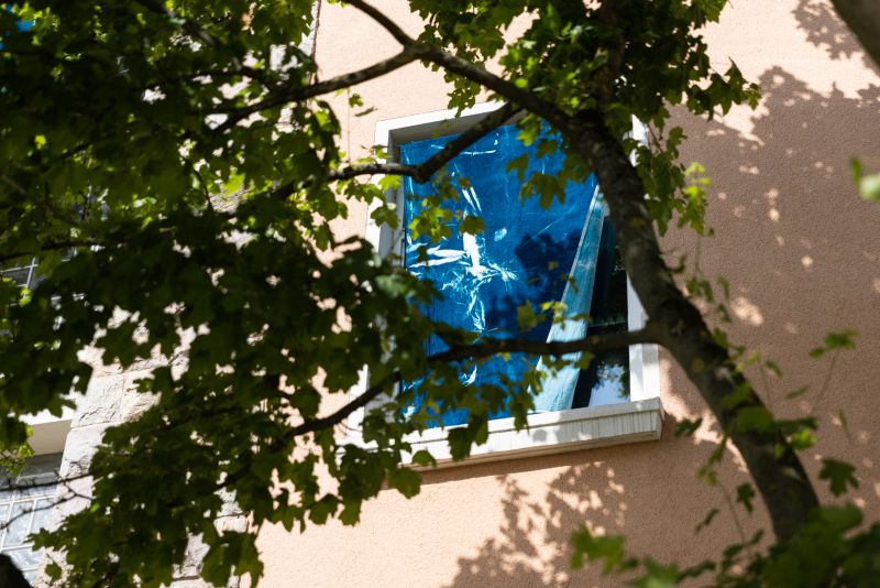 Covered by a large branch in the shade, a window curtained with a blue cyanotype is visible. Sunlight illuminates the window. | photo © Lys Y. Seng